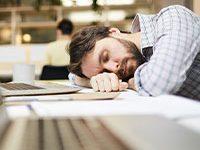 Fotografía de hombre durmiendo sobre un escritorio, al frente tiene una computadora